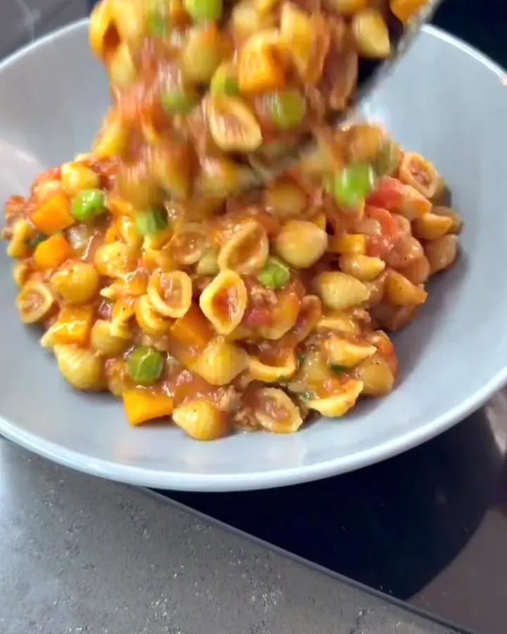 Steaming spoonful of shell pasta with beef, peas, and tomato glaze in a ceramic bowl, topped with Parmesan.
