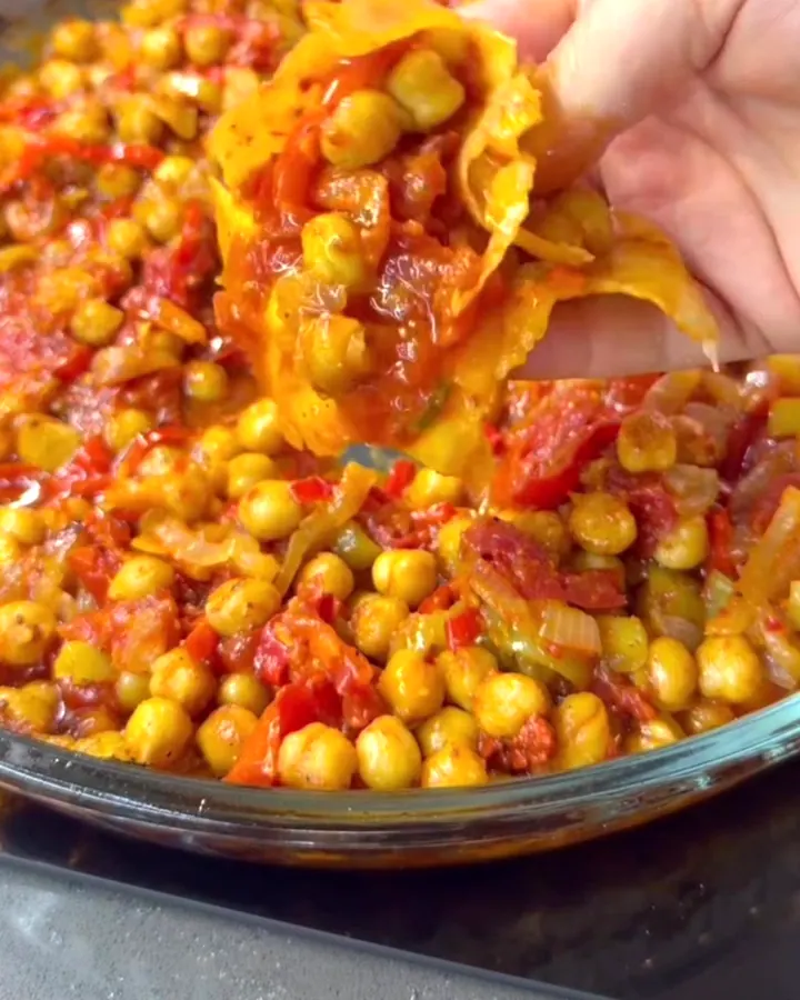 Baked chickpeas with caramelized vegetables in a glass dish, with a piece of flatbread tearing through the mixture.